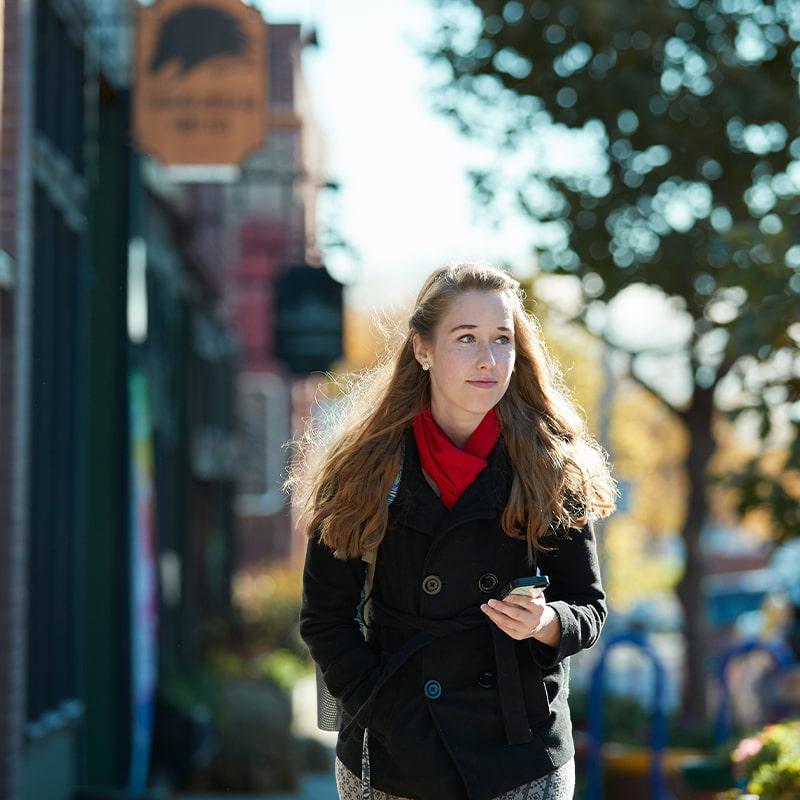 A Drake University student wearing a winter coat and scarf holding a cell phone walking on campus