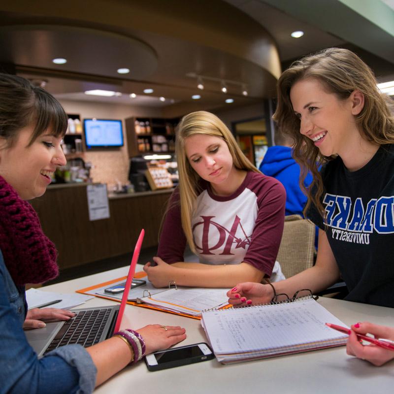 Three Drake University students sitting at a table in a building on campus all working on laptops and notebooks